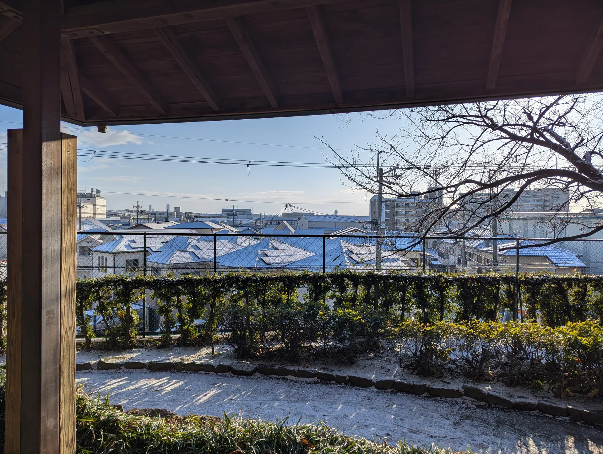 東屋の屋根と柱越しに眺める、雪の残る緑道と街の屋根